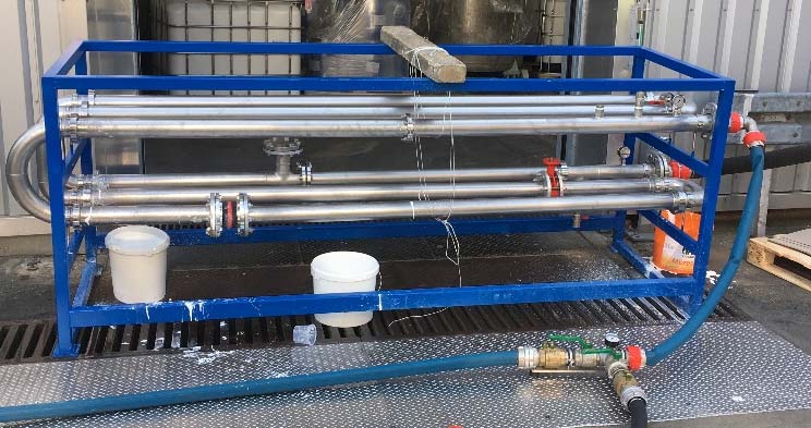 Ein blauer Metallrahmen trägt Edelstahlrohre, Ventile und einen Schlauch, Teil einer Industrieanlage auf einem Gitterboden, die für Spülverfahren und Rohrreinigung verwendet wird.