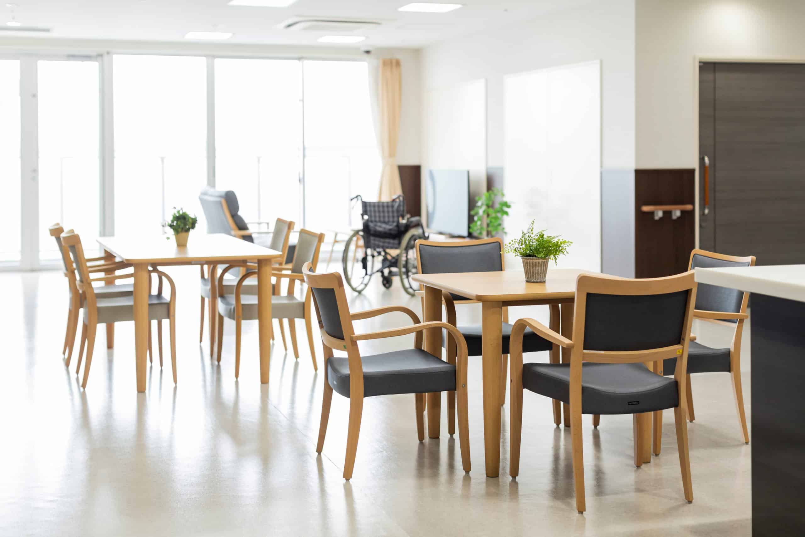 Ein heller, leerer Raum mit Holztischen und -stühlen, Topfpflanzen und einem Rollstuhl neben großen Fenstern strahlt eine ruhige, gemeinschaftliche Atmosphäre aus.