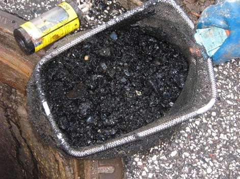 A close-up of a mesh basket filled with dark debris and a tar-like substance, placed next to a yellow can on the ground, likely remnants from pig cleaning.