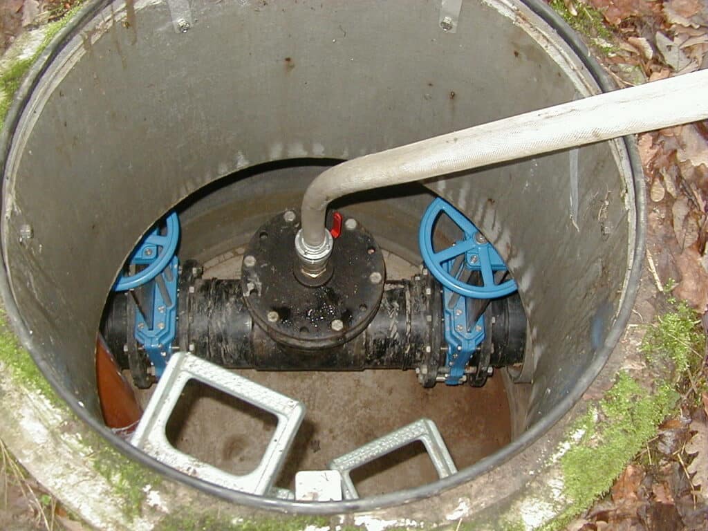 A view inside a cylindrical manhole reveals two blue valve handles and a connected pipe, ready for pig cleaning. The ground surrounding the manhole is covered with leaves.