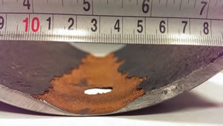 Close-up of a damaged pipe with a rusted hole, positioned under a metal ruler showing a measurement scale from 2 to 8 centimeters, highlighting the need for pig cleaning to maintain pipeline integrity.