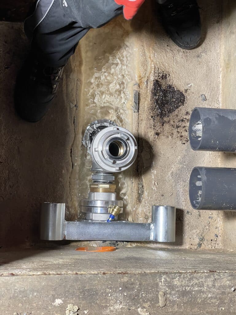 A metal pipe fitting setup in a concrete trench, surrounded by two black hoses used for pig cleaning, with a person standing above, visible from the knees down, wearing black work shoes and pants.