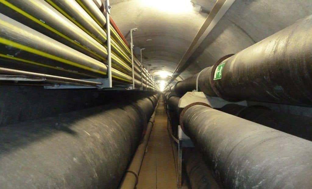 Long underground tunnel with large, insulated pipes and cables running along the ceiling and walls, illuminated by overhead lights.