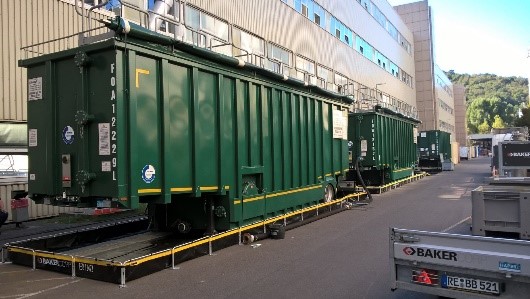 Green industrial containers on ramps near a building suggest the hustle of efficient operations, possibly even pig cleaning processes. The serene trees in the background provide a contrasting backdrop to the scene of organized activity.